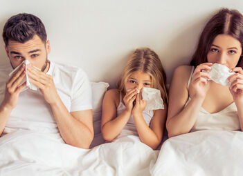 Ein Mann, ein Kind und eine Frau liegen im Bett und schnäuzen sich. Alle schauen dabei in die Kamera.