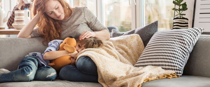 Eine Frau tröstet ihr erkältetes Kind. Beide liegen dabei auf der Couch. 