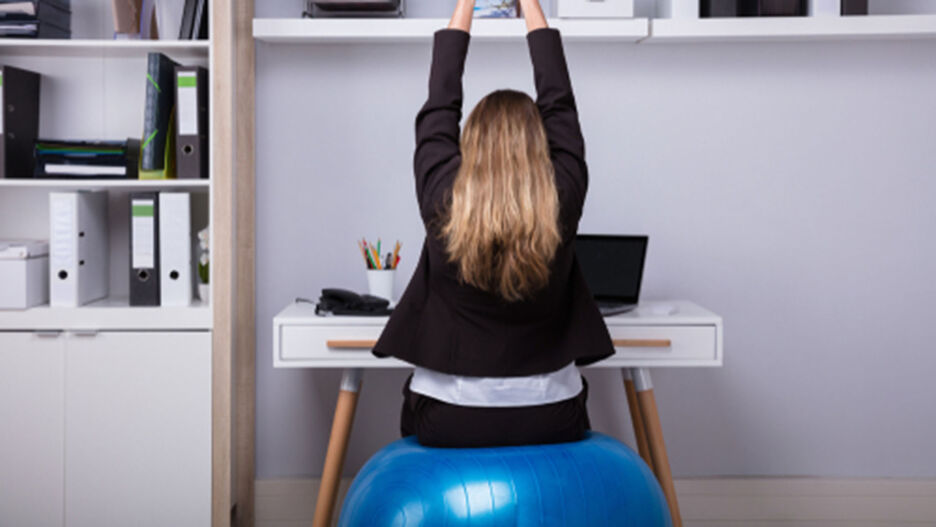 Eine junge Frau sitzt am Schreibtisch auf einem Gymnastikball, um ihren Rücken gleichzeitig zu trainieren und zu entlasten.