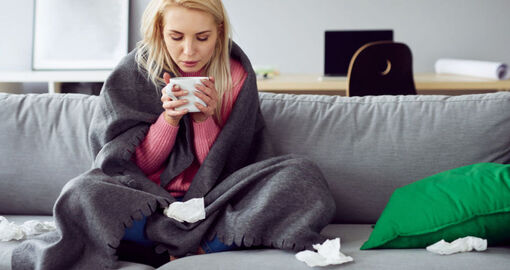 Eine junge Frau sitzt mit Tasse und Decke auf der Couch. 