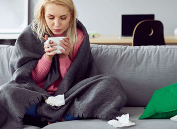 Eine junge Frau sitzt mit Tasse und Decke auf der Couch. 