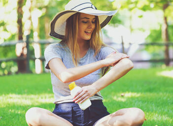 Junge blonde Frau mit weiß-blauem Sommerhut sitzt in kurzer Hose und kurzem T-Shirt im Schneidersitz auf einer grünen Wiese und cremt sich mit Sonnenschutz ein.